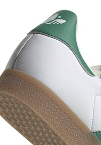 White leather sneaker with a green accent at the heel. Textured gum rubber outsole. Features a logo with three stripes on the heel.