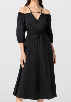 Femme portant une robe midi noire à épaules dénudées, manches trois-quarts et taille froncée, debout devant un fond clair uni.