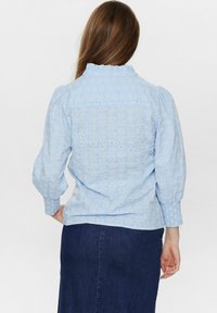 Modra bombažna bluzica z belimi cvetličnimi vezeninami, naborkom okoli vratu in rahlo nabrano manšeto. Obuta je z temnim jeans krilom.