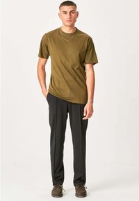 Ung mand står med hænderne i lommerne, iført olivengrøn t-shirt, sorte bukser, brune sko, sølvhalskæde og armbånd.