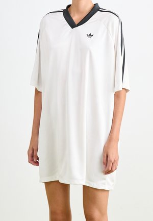 Person iført en løs hvid Adidas-kjole med sort V-hals og sorte striber på korte ærmer mod en ensartet baggrund.