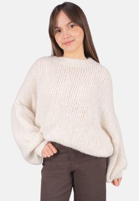 Übergroßer cremefarbener Strickpullover mit strukturiertem Muster, weiten Ballonärmeln und einem gerippten Ausschnitt, kombiniert mit dunkelbraunen Hosen.