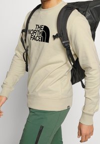 Sudadera beige con un logo negro de "The North Face", puños y dobladillo acanalados; combinada con una mochila negra y pantalones verdes.