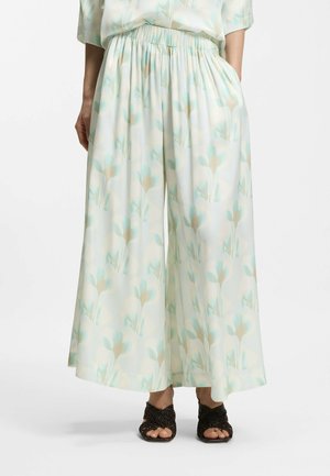 Pantalon crème à jambes larges avec imprimé floral pastel, taille élastique, porté avec des talons ouverts tissés noirs, modèle debout les mains dans les poches.