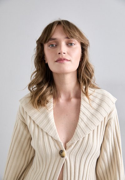 Cream ribbed cardigan with a deep V-neck, wide collar, and a single circular button closure at the front. Features soft, textured fabric.