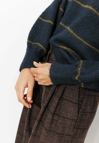 Pull marin bleu avec rayures jaunes, poignets côtelés, associé à une jupe plissée à carreaux marron. Gros plan sur les mains à la taille, mettant en valeur les textures des tissus.