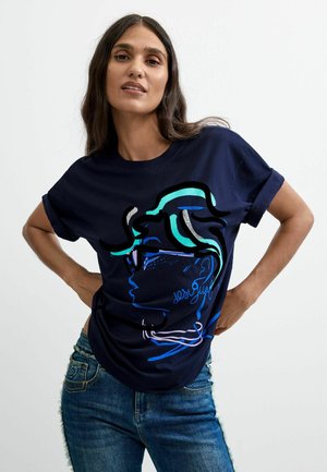 Donna che indossa una maglietta blu navy con un profilo astratto colorato di un volto e jeans blu, con le mani appoggiate sui fianchi.