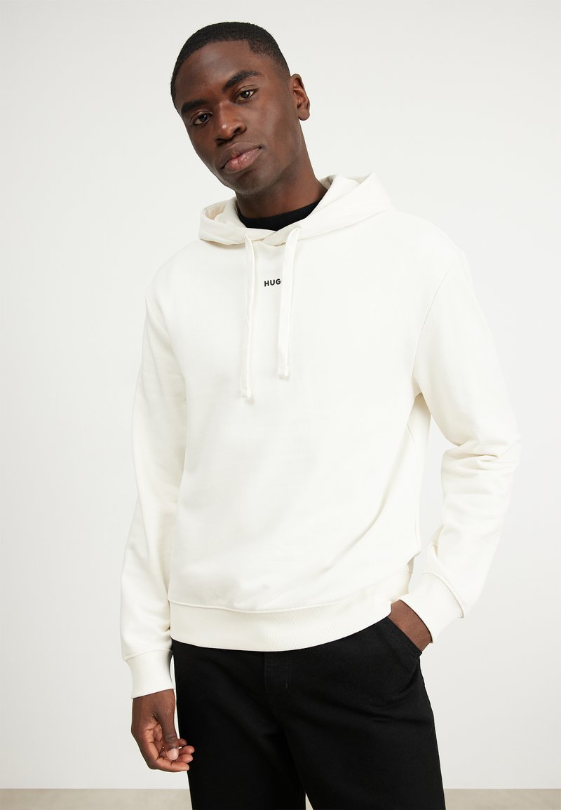 Bijela hoodie sa vezicom, s "HUG" logom na prsima. Mekana tekstura, rebrasti manžeti, uparen s crnim hlačama. Jednostavan dizajn.