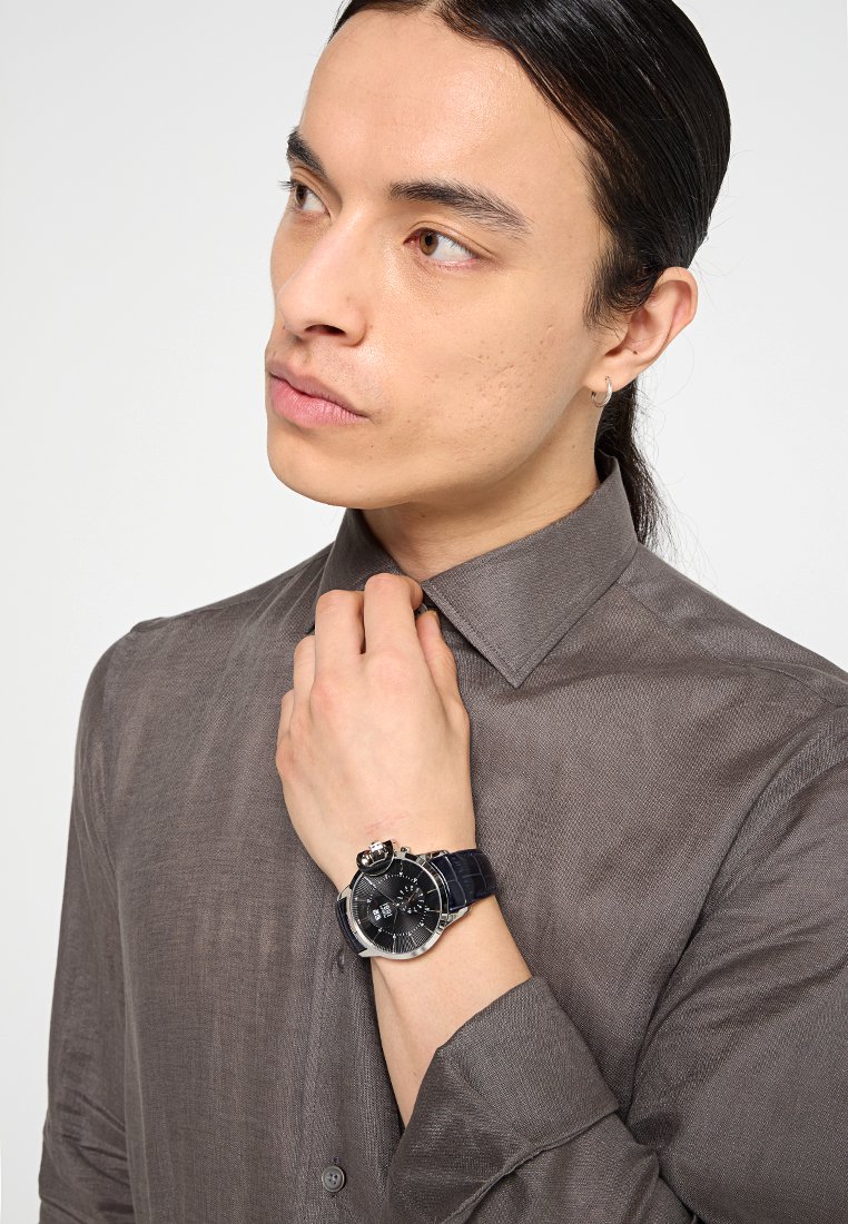 Jeune personne avec des cheveux noirs, portant une chemise à boutons grise et ajustant son col, arborant une montre-bracelet à bracelet en cuir noir au poignet gauche.