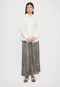 White long-sleeve shirt with a pocket, paired with a pleated, snake-print maxi skirt in shades of gray and black. Black strappy sandals.