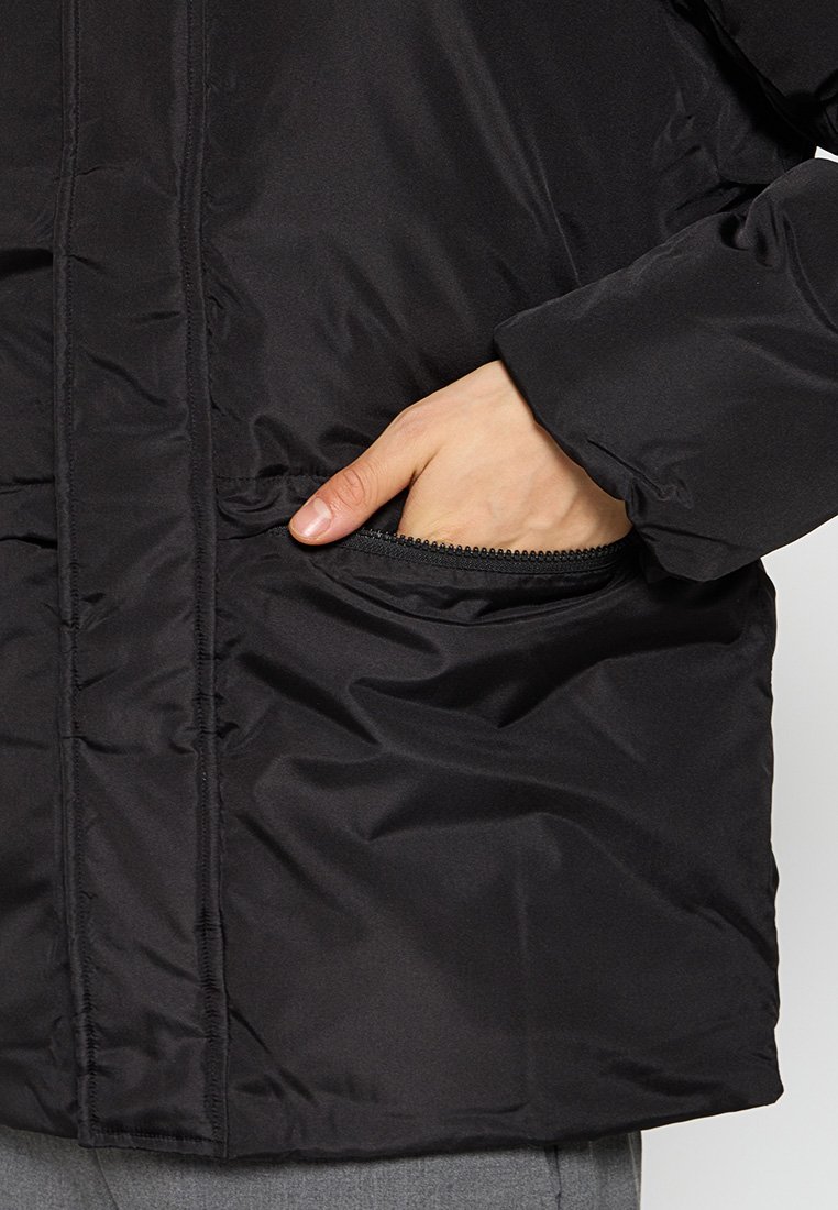 Veste isolante noire avec un extérieur lisse et brillant, dotée d'une poche zippée et d'accents côtelés sur le bord de la poche.