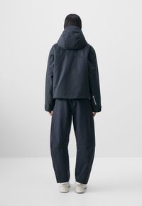 Black, waterproof jacket with a hood, featuring a cropped design and elastic cuffs, paired with loose-fitting black pants and white sneakers.