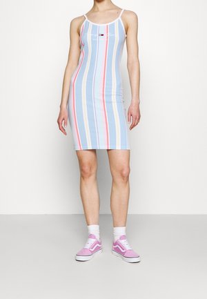 Kobieta w dopasowanej, bezrękawnikowej sukience w pastelowe paski z cienkimi ramiączkami, różowych trampkach i białych skarpetkach, stojąca przy jednolitej ścianie.