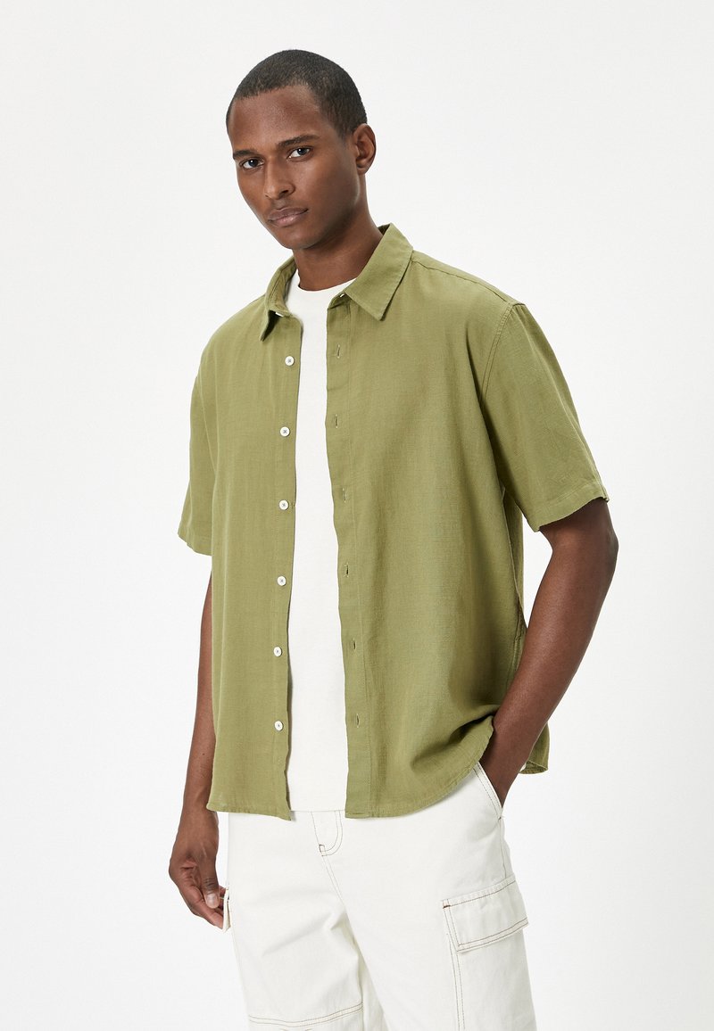 Camisa de manga corta color verde oliva con un cierre de botones en el frente, textura suave y un corte relajado, sobre una camiseta blanca y pantalones de color claro.