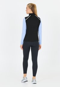 Schwarze ärmellose Weste mit hohem Kragen und reflektierenden Akzenten, über einem hellblauen Langarmshirt getragen, kombiniert mit schwarzen Leggings und silbernen Schuhen.