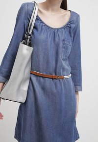 Nainen, jolla on yllään sininen farkkuhaalari, jonka hihat on kääritty, ruskea vyö ja joka kantaa metallinhohtoista hopeista olkalaukkua vasemmalla olkapäällään.