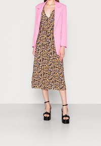 Femme portant un blazer rose, une robe midi noire à fleurs et des sandales à plateforme noires, se tenant devant un fond clair.