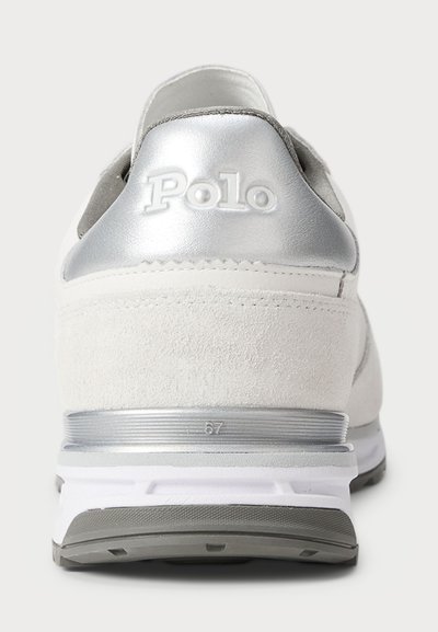 Valge nahast spordijalanõud, millel on hõbedane sünteetiline tagumine osa, esindades embossitud "Polo" logo ja kerget halli tallat, millel on horisontaalsed jooned.