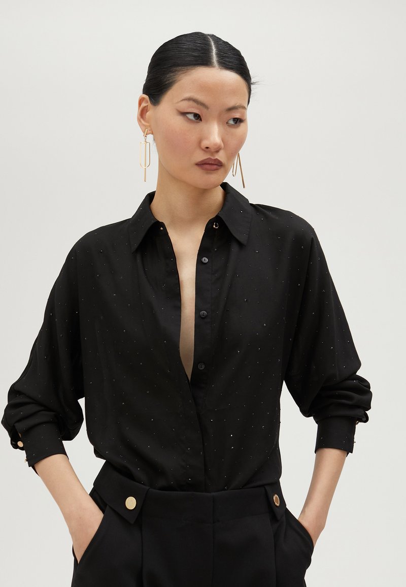 Black shirt made of soft fabric, featuring a loose fit, buttoned cuffs, and small silver dot accents. Trousers with gold button details.