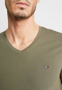 Oliwkowa koszulka v-neck wykonana z miękkiej bawełny, z małym logo w niebieskim, białym i czerwonym kolorze na piersi, o gładkiej fakturze.