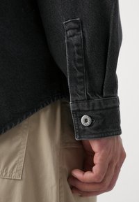 Gros plan de la main d’une personne à côté d’un poignet de chemise en denim noir avec un bouton argenté et d’un pantalon beige avec des coutures visibles.