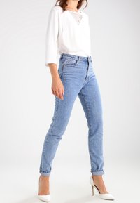 Vit långärmad blus med spetsdetaljer och ljusblå högmidjade jeans, i kombination med vita stilettklackar. Rak bendesign.