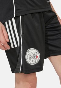 Svarta shorts i andningsbart tyg, med vita ränder på sidorna och en cirkulär logotyp med en detaljerad design på vänster ben.