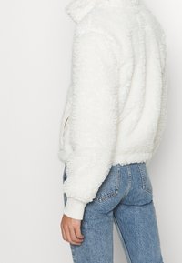 Bílá umělá kožichová bunda s vysokým límcem, žebrovanými manžetami a bočními kapsami, spárovaná se světle modrými džíny s vysokým pasem, s viditelným texturovaným povrchem.