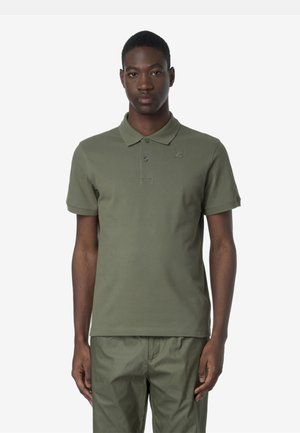 Giovane uomo che indossa una polo verde oliva e pantaloni abbinati, in piedi davanti a uno sfondo bianco semplice, rivolto in avanti con espressione neutra.