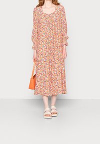 Robe midi florale avec manches bouffantes, fond orange, motifs multicolores. Associée à un sac orange texturé et des sandales à plateforme légères.
