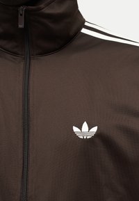 Σκούρο καφέ αθλητικό μπουφάν με φερμουάρ και ψηλό γιακά, λευκό λογότυπο Adidas trefoil στο στήθος και τρεις λευκές ρίγες στον ώμο.
