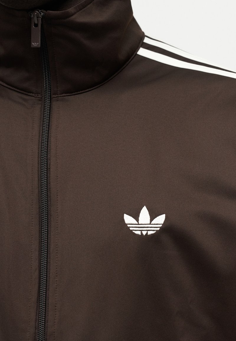 Giacca sportiva marrone scuro con cerniera e collo alto, logo bianco Adidas a trifoglio sul petto, e tre strisce bianche sulla spalla.