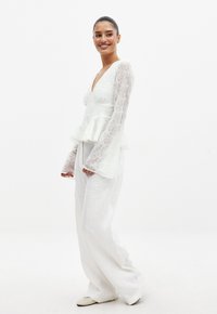 Haut peplum blanc avec un motif en dentelle florale, décolleté en V et longues manches transparentes, associé à un pantalon blanc ample et des chaussures de couleur claire.