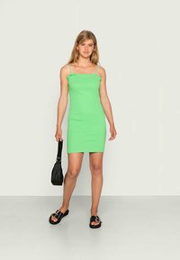 Vestido verde justo com alças finas, decote franzido e uma textura suave, combinado com sandálias pretas e uma pequena mala preta.