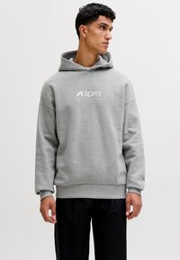 Nuori mies, joka pukeutunut harmaaseen huppariin, jossa teksti "aprel" ja koordinaatit, katsoo sivulle, seisoo yksinkertaisen valkoisen taustan edessä.