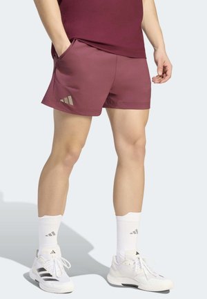 Mand iført bordeaux atletikshorts, hvide ankelsokker og hvide Adidas-sneakers står med den ene hånd i lommen.