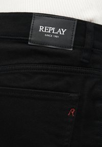 Μαύρο τζιν με κεντημένη μαύρη ετικέτα που γράφει "REPLAY SINCE 1981" πάνω από την πίσω τσέπη και μικρή κόκκινη κέντηση "R".