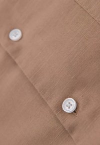 Tissu marron avec une texture en lin, comportant deux boutons blancs. Le tissu présente un tissage subtil et des détails de couture visibles.