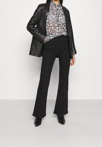 Personne portant un blazer en cuir noir par-dessus un chemisier floral, un pantalon évasé taille haute noir et des bottines noires à bout pointu.