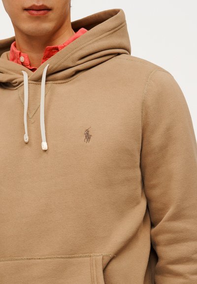 Άνδρας που φοράει μπεζ φούτερ με λευκά κορδόνια και μικρό κεντημένο λογότυπο, από πάνω από κόκκινο πουκάμισο με κουμπιά, σε λευκό φόντο.