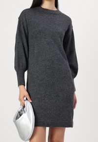 Robe en maille grise à manches longues, col rond et poignets côtelés. Le mannequin tient une pochette argentée. Design simple, texture douce.