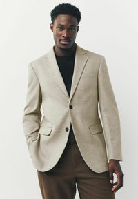 Blazer beige texturizado con dos bolsillos frontales, solapas en pico y botones oscuros, llevado sobre una camisa negra y pantalones marrones a medida.