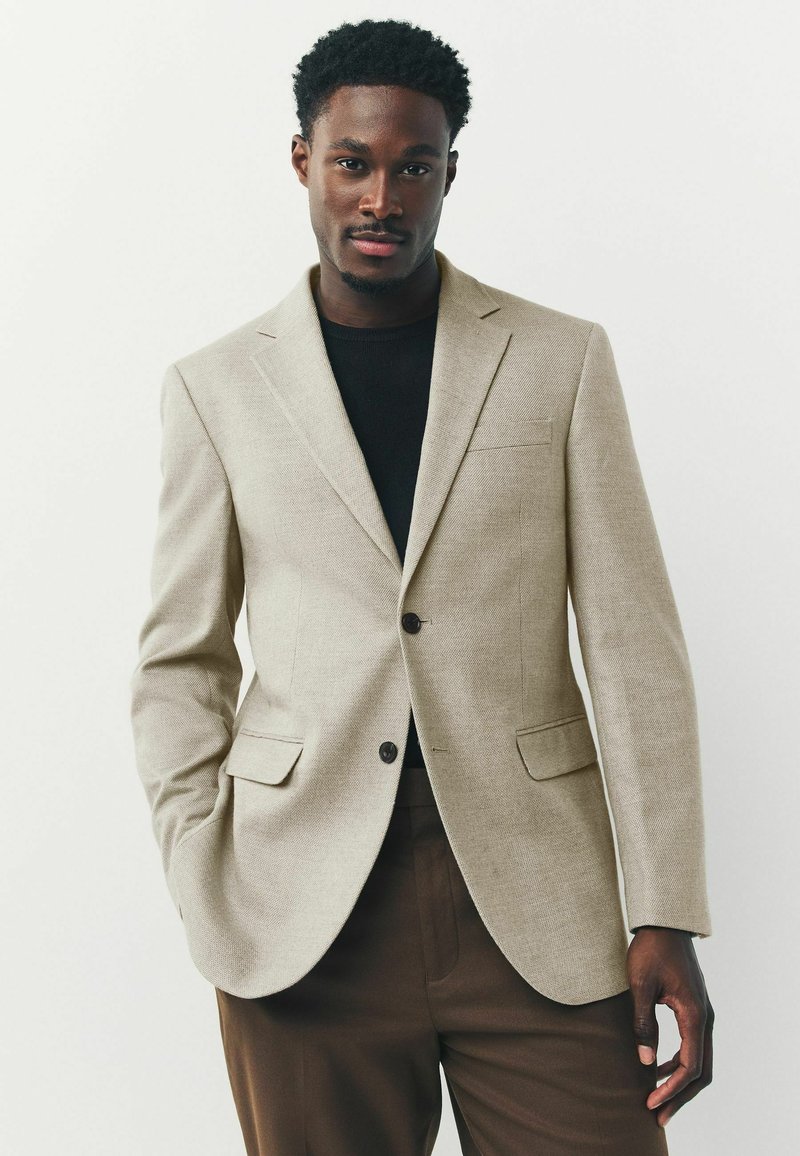 Beige strukturierter Blazer mit zwei Vorderjackentaschen, Reverskragen und dunklen Knöpfen, getragen über einem schwarzen Hemd mit braunen maßgeschneiderten Hosen.