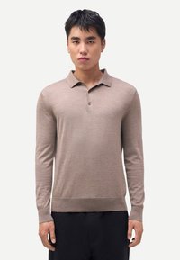 Junger Mann trägt ein beiges Langarm-Poloshirt und schwarze Hose, steht vor einem einfarbigen hellgrauen Hintergrund.