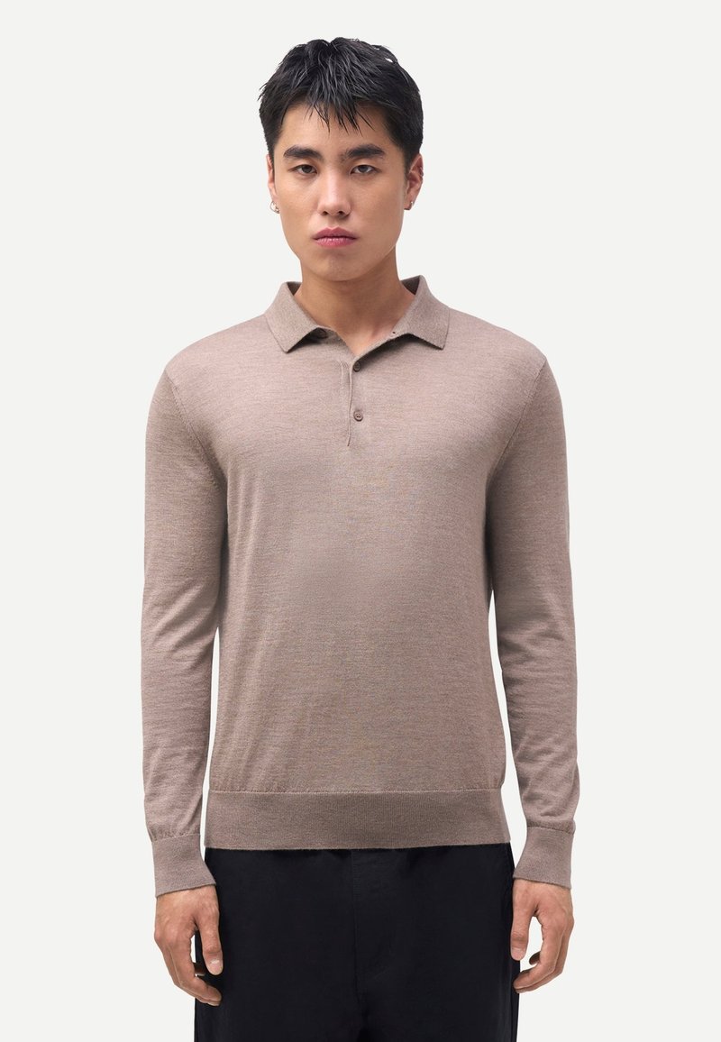 Junger Mann trägt ein beiges Langarm-Poloshirt und schwarze Hose, steht vor einem einfarbigen hellgrauen Hintergrund.