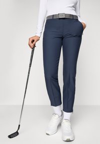 Persoană îmbrăcată cu pantaloni bleumarin, bluză albă cu mâneci lungi, adidași albi, ținând o crosă de golf, stând în fața unui fundal simplu.