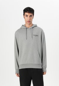 Felpa grigia realizzata in tessuto morbido, con tasca a canguro frontale, cappuccio con lacci e testo del logo nero sul petto. Indossata con pantaloni neri.