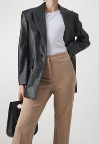 Blazer en cuir synthétique noir avec un motif crocodile, haut blanc ajusté, et pantalon beige à jambes larges. Sac à main noir avec une anse découpée.