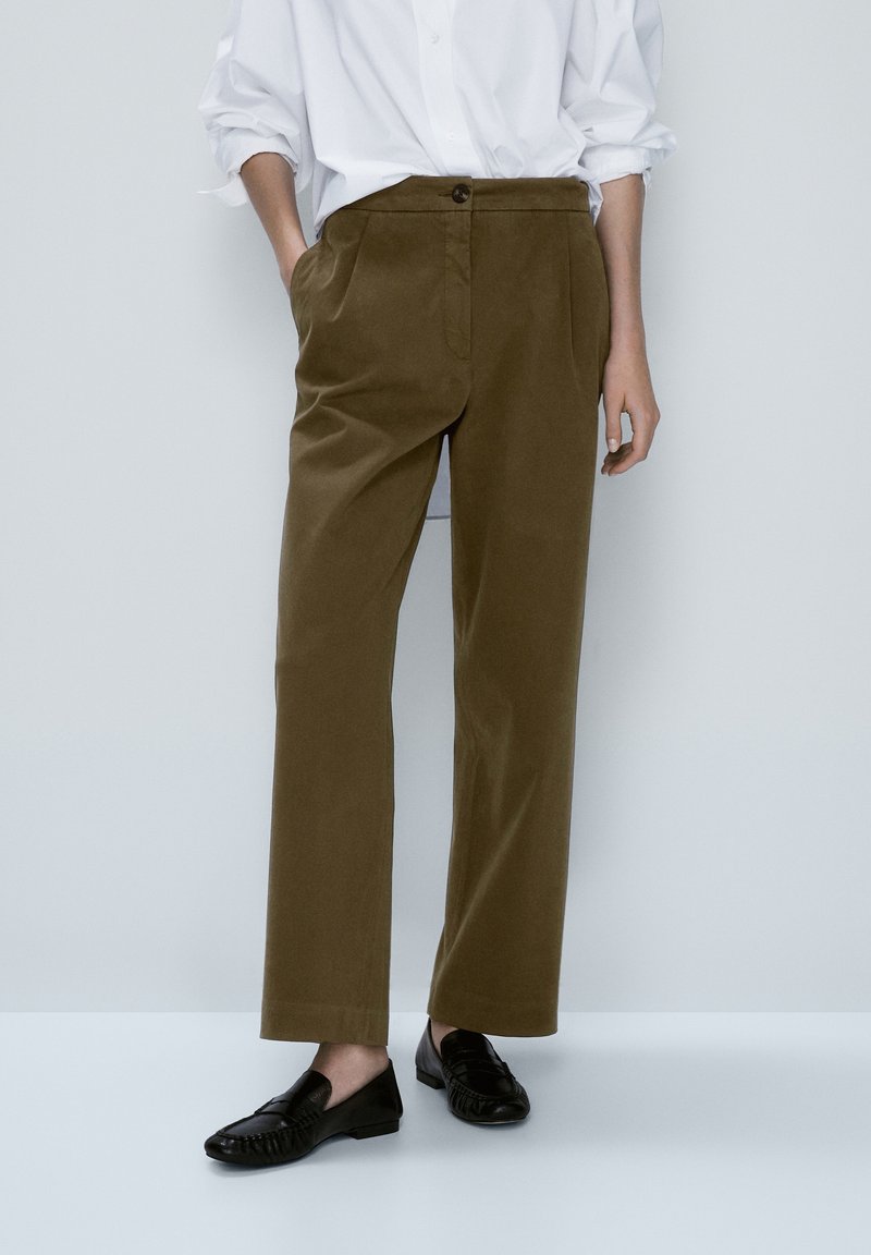 Personne portant un pantalon vert olive à jambes droites, une chemise blanche à boutons avec les manches retroussées, et des mocassins noirs, debout avec une main dans la poche.