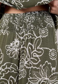 Tessuto verde oliva con stampa floreale bianca, dotato di una vita arricciata. La texture appare morbida e leggera, con un design fluido.